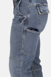 Side view of a woman wearing Dovetail Workwear's Day Construct Work Pant in Vintage Blue denim, showcasing the reinforced front pocket with a phone visible and reinforced stitching details
