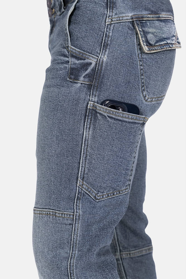 Side view of a woman wearing Dovetail Workwear's Day Construct Work Pant in Vintage Blue denim, showcasing the reinforced front pocket with a phone visible and reinforced stitching details