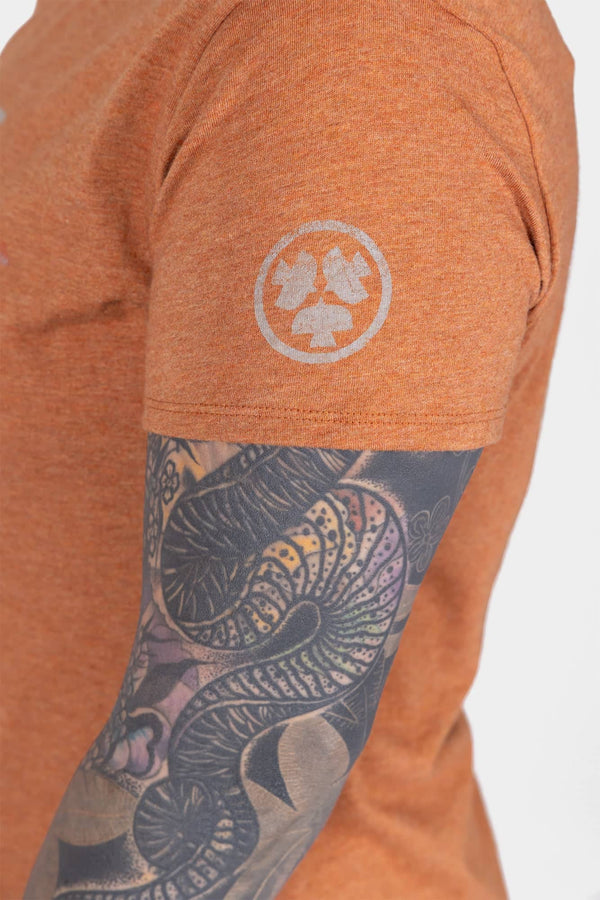 Side view of a woman wearing the Dovetail Workwear GO TO™ "Get Dirty" Graphic Work Tee in rust orange with graphic logo, paired with dark patterned sleeve tattoo