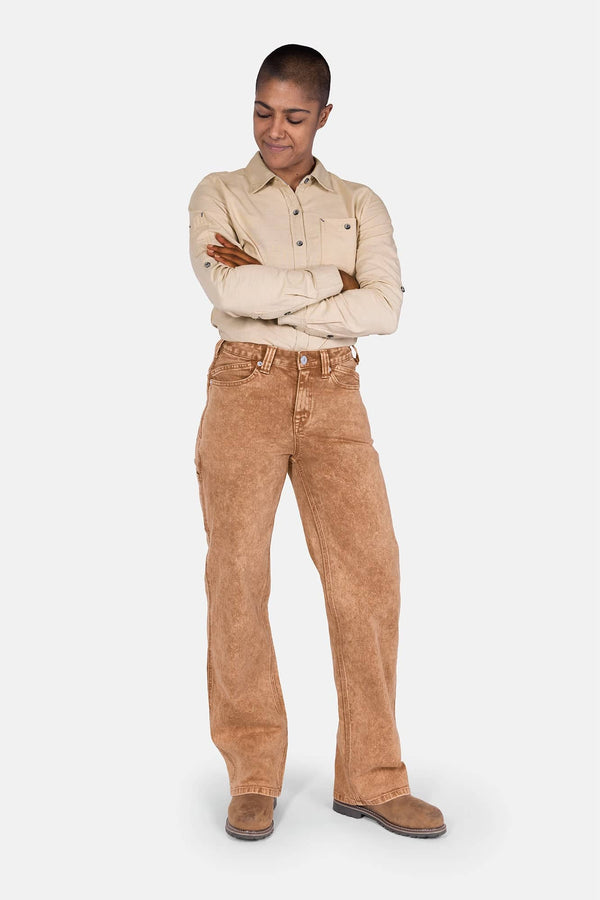 Woman wearing Dovetail Workwear's Old School Wide Leg Mineral Wash Jeans in Sunbaked Copper with a beige button-up shirt, arms crossed, looking down
