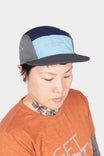 Woman wearing the Ocean Blue Dovetail Workwear Pack Cap, a 5-panel cap with light blue front panel, dark navy top, gray mesh sides, and black brim, paired with an orange t-shirt