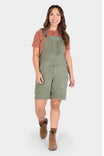 Dovetail Workwear Utility Short Overalls in Light Kelp Green worn by a woman with a rust t-shirt and brown boots, featuring front bib pocket with snap closure and flex suspenders
