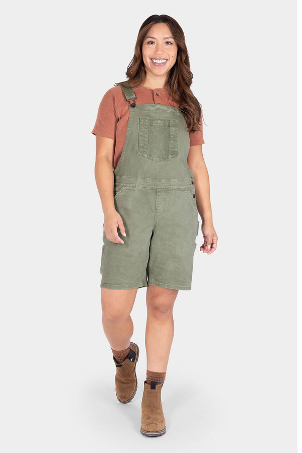 Dovetail Workwear Utility Short Overalls in Light Kelp Green worn by a woman with a rust t-shirt and brown boots, featuring front bib pocket with snap closure and flex suspenders