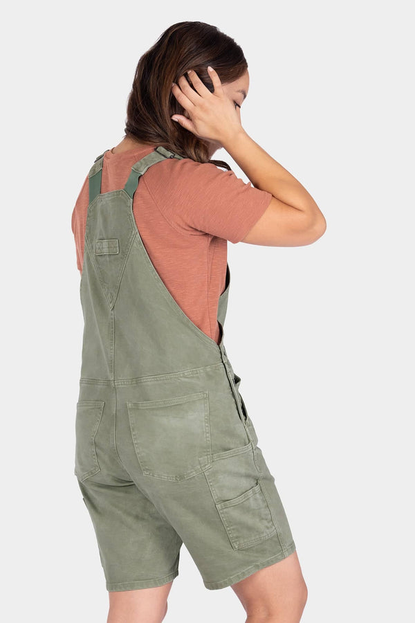 Back view of a woman wearing Dovetail Workwear's Utility Short Overalls in Light Kelp Green, showing the bib pocket, tool loops, and relaxed fit design