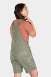Back view of a woman wearing Dovetail Workwear Utility Short Overalls in Light Kelp Green over a rust-colored shirt, showing the bib pocket and relaxed fit design