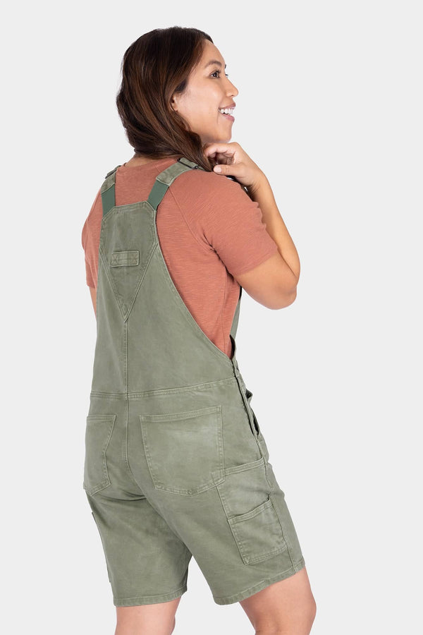 Back view of a woman wearing Dovetail Workwear Utility Short Overalls in Light Kelp Green over a rust-colored shirt, showing the bib pocket and relaxed fit design