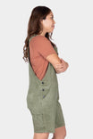 Side view of a woman wearing Dovetail Workwear's Utility Short Overalls in Light Kelp Green, showing the relaxed fit, bib pocket, and tool loop with a coral t-shirt underneath