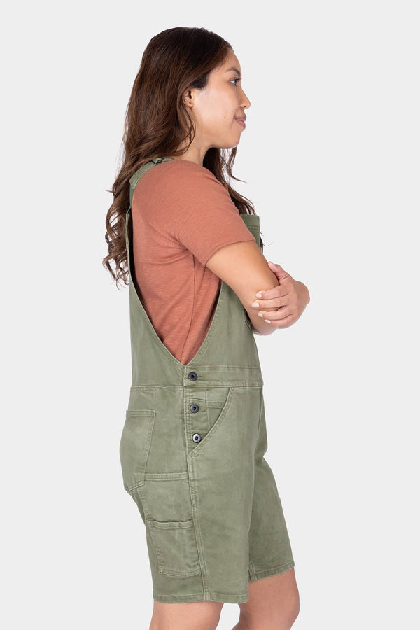 Side view of a woman wearing Dovetail Workwear's Utility Short Overalls in Light Kelp Green, showing the relaxed fit, bib pocket, and tool loop with a coral t-shirt underneath
