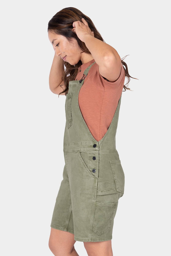 Side view of a woman wearing Dovetail Workwear's Utility Short Overalls in Light Kelp Green with a coral tank top, featuring visible bib pocket and hip clip