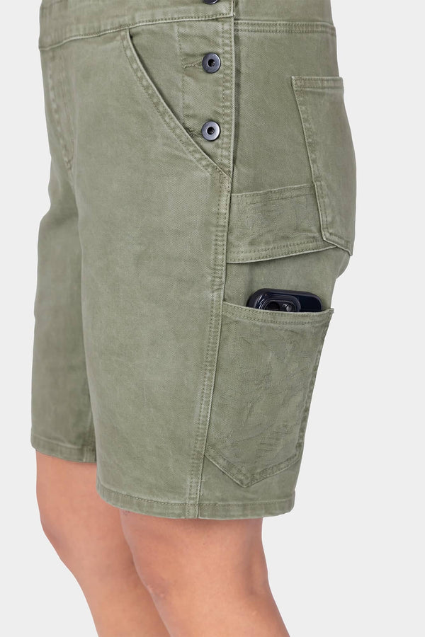Side view of a woman wearing Dovetail Workwear's Utility Short Overalls in Light Kelp Green, featuring multiple cargo pockets and a phone pocket with visible snap buttons