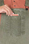 Detail view of a woman's hand retrieving a Bicycle card from the bib pocket of Dovetail Workwear's Utility Short Overalls in Light Kelp Green, showcasing the snap closure pocket feature