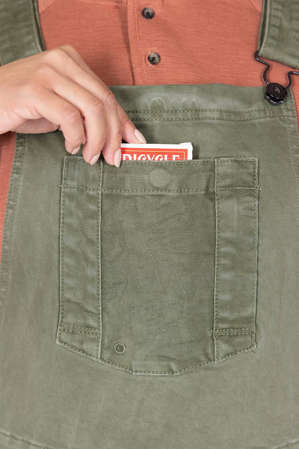 Detail view of a woman's hand retrieving a Bicycle card from the bib pocket of Dovetail Workwear's Utility Short Overalls in Light Kelp Green, showcasing the snap closure pocket feature