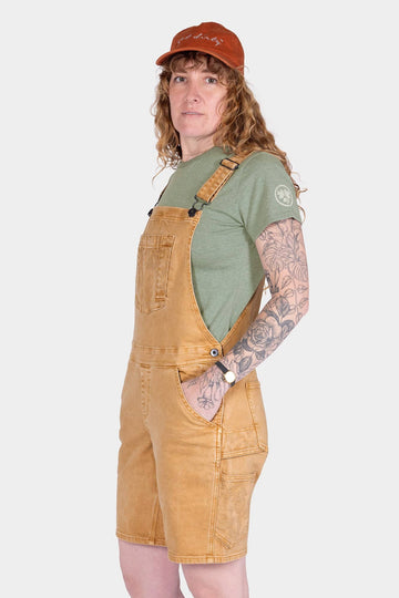 Side view of a woman wearing Dovetail Workwear's Utility Short Overalls in Tan Sandstone, featuring flex suspenders, multiple pockets, and a relaxed fit over a green t-shirt