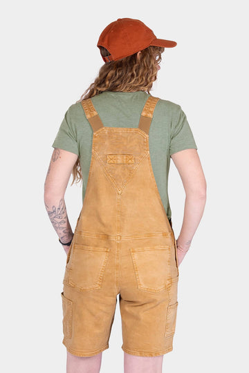 Back view of a woman wearing Dovetail Workwear's Utility Short Overalls in Tan Sandstone, showcasing the bib pocket, dual rear pockets, and adjustable flex suspenders