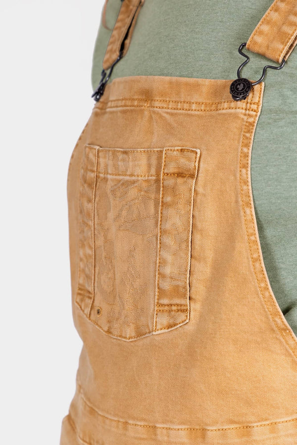 Detail view of the Tan Sandstone Utility Short Overalls by Dovetail Workwear, showing the bib pocket with snap closure and Hip Clip on a woman wearing a sage green shirt