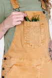 Woman wearing Dovetail Workwear Utility Short Overalls in Tan Sandstone, displaying the bib pocket and adjustable flex suspenders