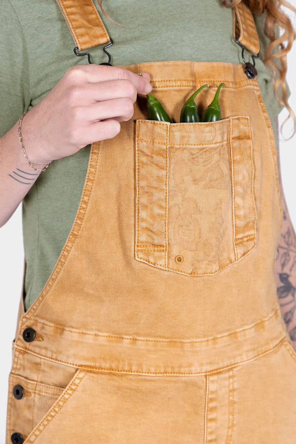 Woman wearing Dovetail Workwear Utility Short Overalls in Tan Sandstone, displaying the bib pocket and adjustable flex suspenders