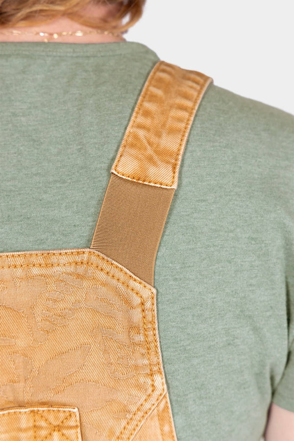 Back view of a woman wearing Dovetail Workwear's Utility Short Overalls in Light Kelp Green, showing flex suspenders and jacquard texture details