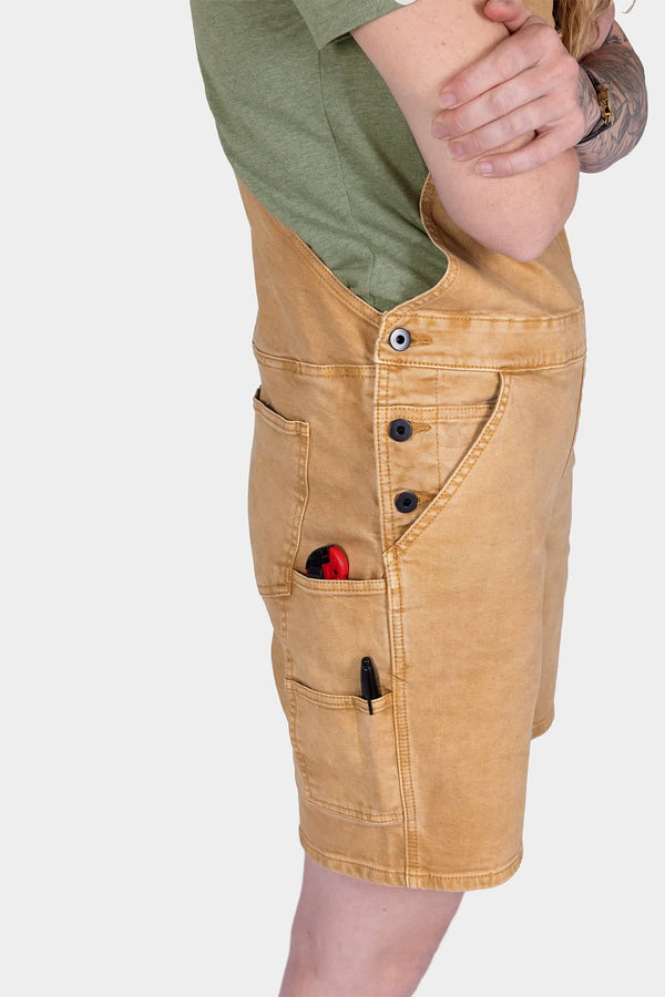 Side view of a woman wearing Dovetail Workwear Utility Short Overalls in Tan Sandstone, displaying multiple pockets, snap closures, and tool loops