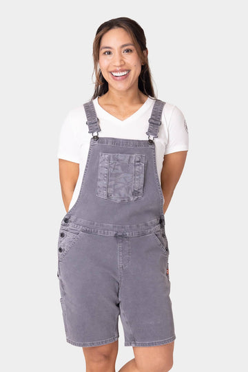 Woman smiling while wearing Dovetail Workwear Utility Short Overalls in Driftwood Grey with white t-shirt, featuring bib pocket, suspenders, and multiple functional pockets