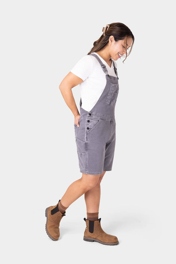 Side view of a woman wearing Dovetail Workwear's Utility Short Overalls in Driftwood Grey, showcasing the relaxed fit, bib pocket, and tool loop details with brown boots