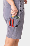 Side view of a woman wearing Dovetail Workwear Utility Short Overalls in Driftwood Grey, displaying multiple pockets including nested side pockets holding red pliers and a small melon