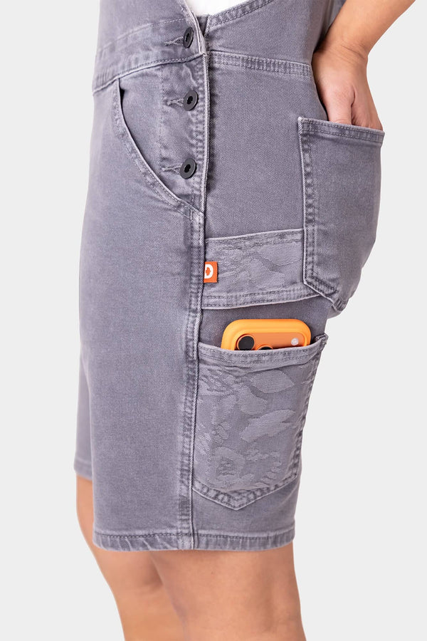Side view of a woman wearing Dovetail Workwear's Utility Short Overalls in Driftwood Grey, showcasing multiple pockets including a phone pocket with an orange device visible