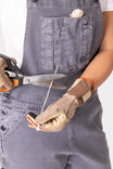 Woman wearing Dovetail Workwear Utility Short Overalls in Driftwood Grey displaying work gloves and scissors, featuring visible bib pocket with snap closure and tool loop details