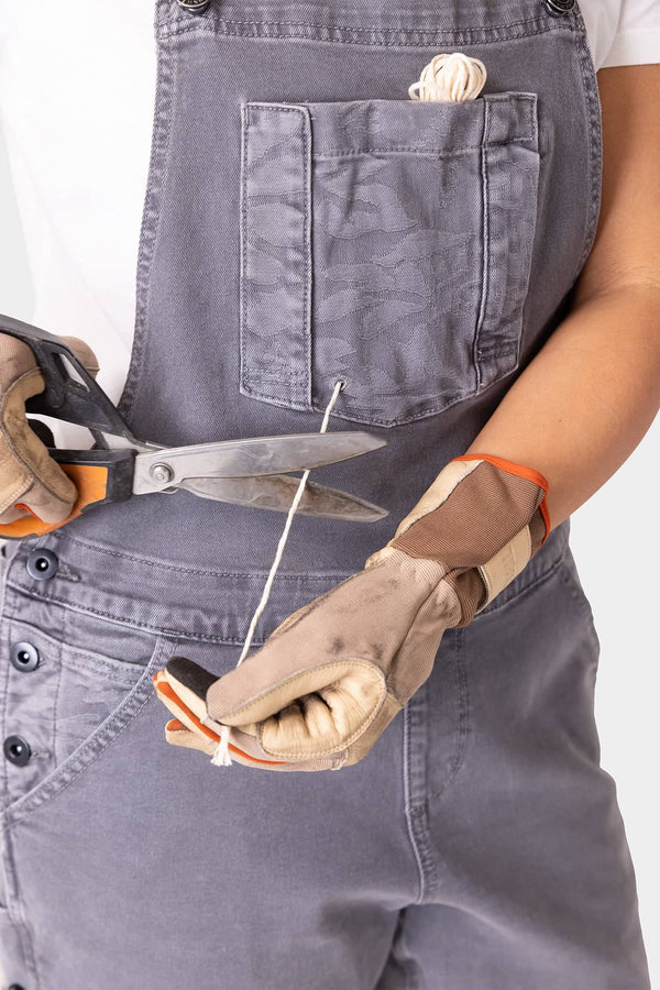 Woman wearing Dovetail Workwear Utility Short Overalls in Driftwood Grey displaying work gloves and scissors, featuring visible bib pocket with snap closure and tool loop details