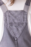 Back view of Dovetail Workwear Utility Short Overalls in Driftwood Grey, showing bib pocket with snap closure, tool loop, and flex suspenders on a woman