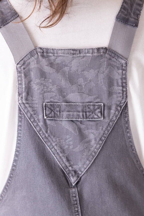 Back view of Dovetail Workwear Utility Short Overalls in Driftwood Grey, showing bib pocket with snap closure, tool loop, and flex suspenders on a woman