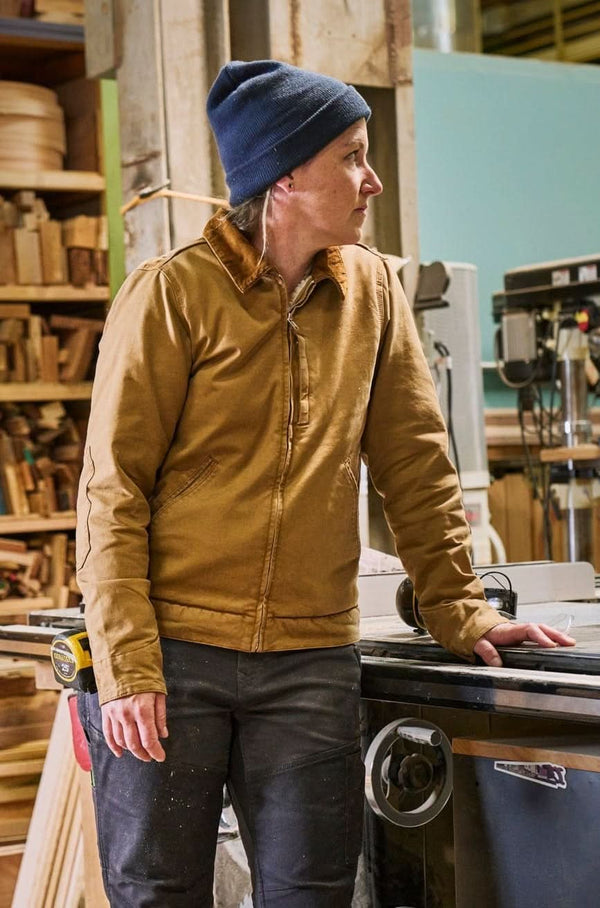 Woman wearing the Dovetail Workwear Old School Trucker Canvas Work Jacket in Vintage Brown with a blue beanie, standing in a workshop next to machinery
