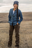 Woman wearing Dovetail Workwear Day Construct Pant in Brown Canvas with denim shirt and beanie, standing in an open field