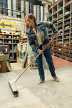 Woman wearing the Loden Green Plaid Durham Reversible Quilted Shirt Jac from Dovetail Workwear while sweeping in a warehouse, showcasing the flannel side of the reversible jacket