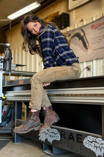 Woman sitting on workbench wearing Dovetail Workwear Old School High Rise Work Pant in Beechwood Brown denim with blue plaid shirt and brown lace-up boots