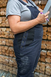 Pregnant woman wearing Dovetail Workwear Freshley Maternity Overalls in Navy Stretch Canvas, holding a clipboard against a brick wall background