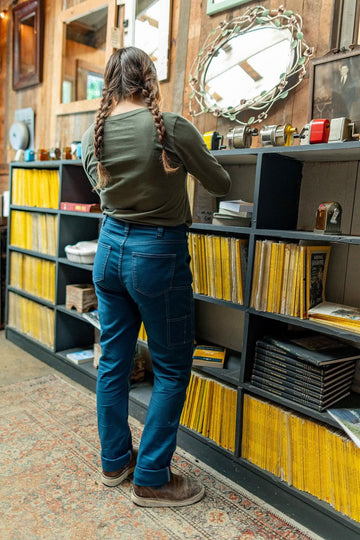 Back view of a woman wearing Dovetail Workwear's GO TO™ Stretch Canvas Pant in Atlantic Blue, showcasing the straight leg fit and nested pockets while organizing shelves in a workshop