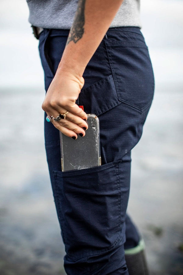 Side view of a woman wearing Dovetail Workwear's Britt X Ultralight Work Pant in Navy Ripstop, showing the cargo-style pocket and Hip Slot™ detail near water
