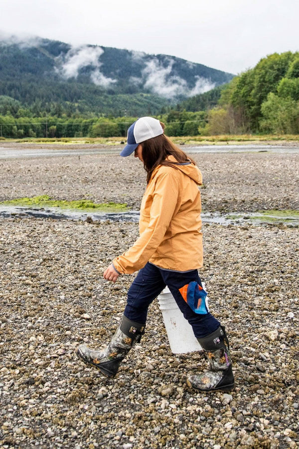 Side view of a woman wearing Dovetail Workwear's Britt X Ultralight Work Pant in Navy Ripstop, running on a rocky beach with mountains in the background