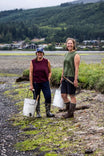 Two women wearing Dovetail Workwear Bellows Work Tank Tops in Blade Green and Elderberry (Maroon) while doing outdoor work near water with tools