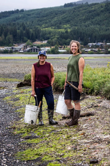 Two women wearing Dovetail Workwear Bellows Work Tank Tops in Blade Green and Elderberry (Maroon) while doing outdoor work near water with tools