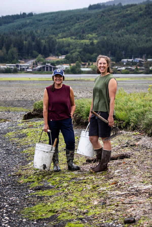 Two women wearing Dovetail Workwear Bellows Work Tank Tops in Blade Green and Elderberry (Maroon) while doing outdoor work near water with tools