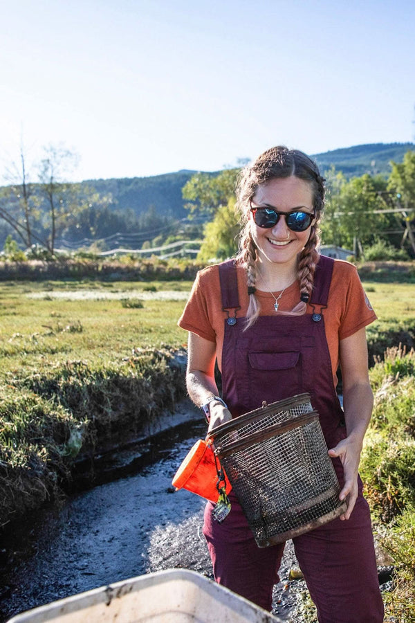 Woman wearing Elderberry Maroon Freshley Overalls in Ultralight Ripstop by Dovetail Workwear while holding a harvest basket by a stream in a mountainous valley