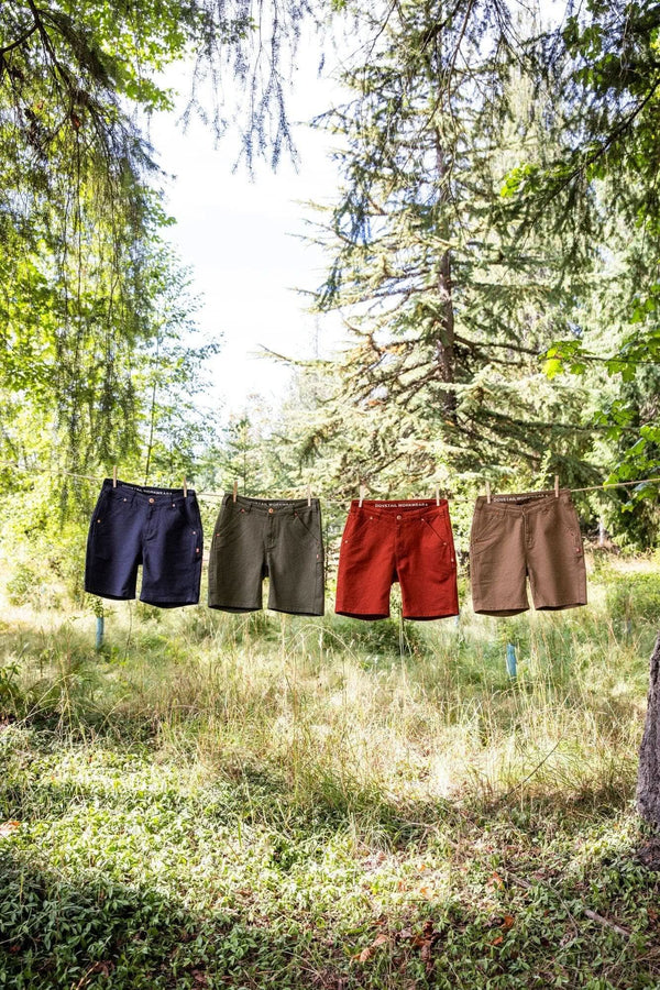 Four Dovetail Workwear GO TO™ Stretch Canvas Shorts in cement grey, kelp green, paprika, and sawdust brown hang on a clothesline in a forest setting