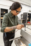 Woman wearing Dovetail Workwear's Givens Stretch Chamois Work Shirt in Patina Green, demonstrating the pencil pocket on the sleeve while working in a shop