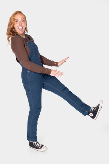 Side view of a woman wearing Dovetail Workwear GO TO™ Stretch Canvas Overalls in Atlantic Blue with flex suspenders, demonstrating the flattering fit and articulated knee design