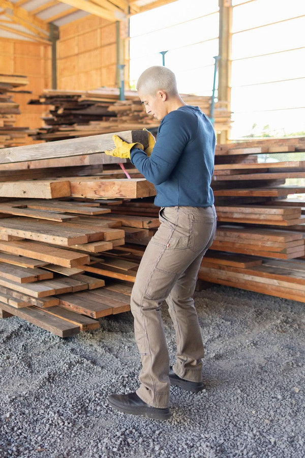 Woman wearing Dovetail Workwear's Honed Long Sleeve Thermal Henley in Atlantic Blue, inspecting lumber at a worksite, showcasing the waffle knit thermal base layer and reinforced elbows.