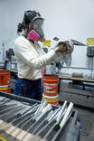 Woman wearing Dovetail Workwear's Honed Long Sleeve Thermal Henley in Oatmeal with waffle knit thermal base layer and reinforced elbows, demonstrating its durability in an industrial workshop setting