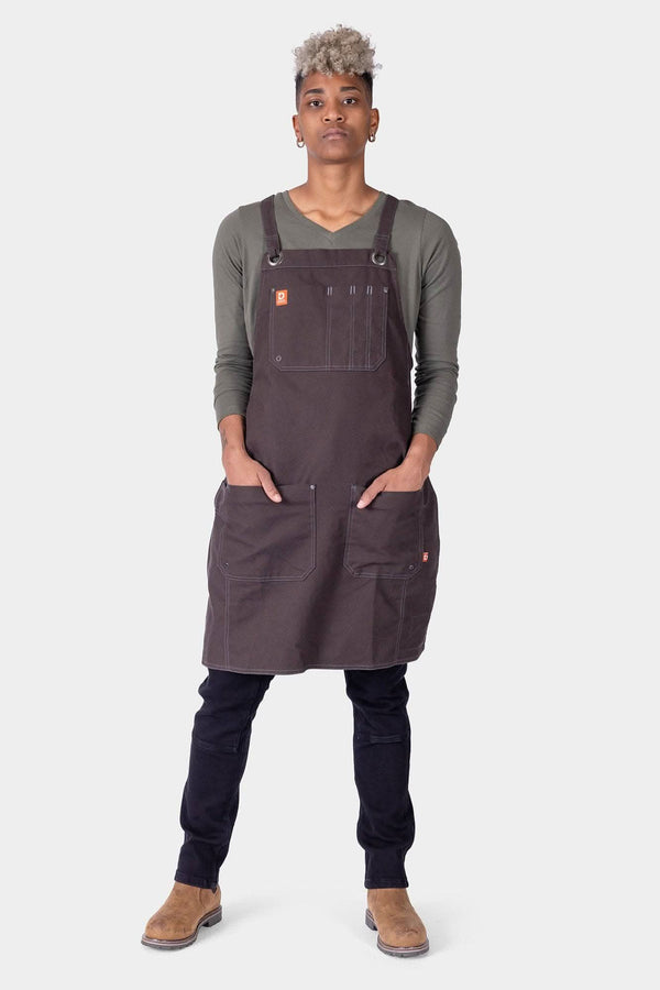 Woman wearing the Dovetail Workwear Obana Work Apron in Kodiak Brown with cross-back straps, displaying multiple pockets and tool organization features