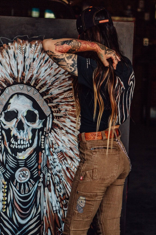 Back view of a woman wearing Dovetail Workwear Rae Ripple X NoSo Patch in tan cargo pants, displaying peel-and-stick patches on denim and artwork beside her